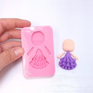 Könnte beinhalten: Eine rosa Silikonform in Form einer Puppe mit Rüschenrock, gehalten von einer Hand. Daneben eine Puppe aus Ton mit lila Rüschenrock und leerem Kopf und Armen.