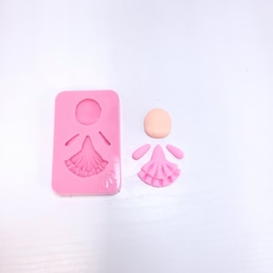 Könnte beinhalten: Eine rosa Silikonform und Puppenteile. Die Form ist rechteckig mit einer Puppenform. Die Puppenteile umfassen einen pfirsichfarbenen Kopf, rosa Arme und ein rosa Kleid. Das Puppenkleid hat ein Rüschendesign.