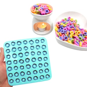 Puede incluir: Un molde de silicona turquesa con numerosas cavidades circulares, junto a un quemador de aceite de cerámica blanca con una vela encendida y un plato en forma de corazón lleno de ceras aromáticas coloridas en forma de anillo.