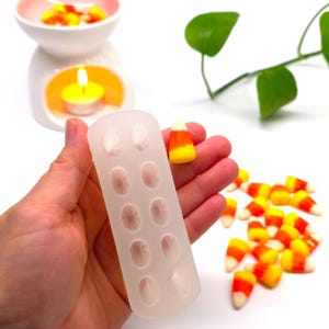 May include: A translucent silicone mould with ten oval-shaped cavities, held alongside a single candy corn piece. The candy corn features a white tip, orange centre, and yellow base. Additional candy corn pieces are scattered nearby.