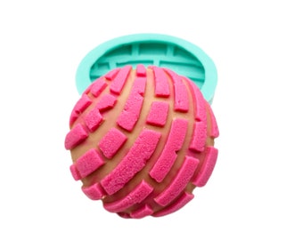 Puede incluir: Un pan concha rosa con un patrón texturizado, en un molde de silicona turquesa. El pan tiene una base marrón claro y está cubierto con un diseño rosa vibrante. La imagen está sobre un fondo blanco.