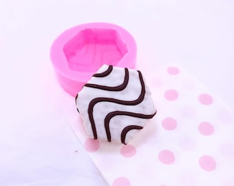 May include: A pink silicone mold shaped like a hexagon, with a white and black striped cake inside. The cake is sitting on a white napkin with pink polka dots.