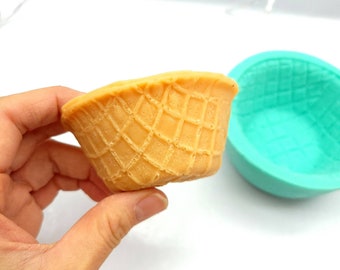 Puede incluir: Un molde de cono de waffle de silicona, de color turquesa. El molde está sobre una superficie blanca. Al lado, un cono de waffle, sostenido en una mano, de color marrón claro.