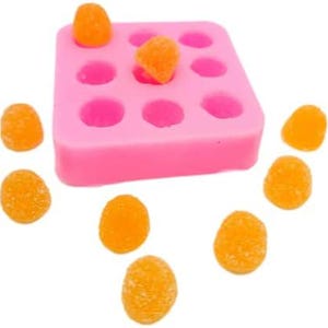 May include: A pink silicone mould with nine circular cavities, holding three orange sweets. Scattered around the mould are additional orange sweets. The sweets have a textured, sugary coating. The image is set against a white background.