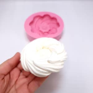 Op de afbeelding: Een witte, gewervelde lekkernij wordt in een hand gehouden, naast een roze siliconen mal. De ronde mal heeft een bloemenontwerp. De lekkernij lijkt een gebakken product te zijn, mogelijk meringue of glazuur, met een getextureerd, gelaagd uiterlijk.