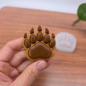 Peut inclure: Un objet en forme de patte d'ours marron avec une finition lisse et brillante, doté de cinq coussinets distincts avec des griffes. L'objet présente une surface texturée et est accompagné d'un moule en silicone translucide et souple.