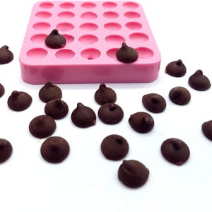 May include: A pink silicone mould with numerous circular cavities, some filled with dark chocolate chips. Several chocolate chips are scattered around the mould on a white surface. The chips are teardrop-shaped.