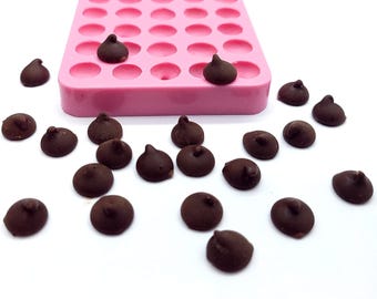 May include: A pink silicone mold with multiple round cavities, partially filled with dark chocolate chips. Several chocolate chips are scattered around the mold on a white surface. The chips have a teardrop shape.
