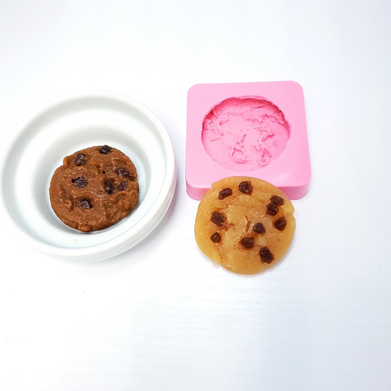 May include: A chocolate chip cookie next to a white bowl and a pink silicone mold. The cookie is brown with dark chocolate chips. The mold is square with a cookie-shaped indentation.
