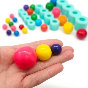 Puede incluir: Tres gominolas redondas y de colores en diferentes tamaños. Las gominolas son de color rosa, amarillo y morado. Las gominolas están en una mano.