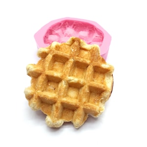 May include: A close-up of a golden brown waffle, with a pink silicone mould in the background. The waffle has a square grid pattern and is slightly puffed up.