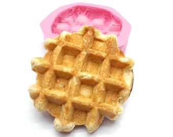 May include: A close-up of a golden brown waffle, with a pink silicone mold in the background. The waffle has a square grid pattern and is slightly puffed up.