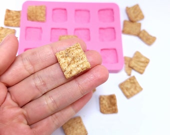 May include: A hand holding a square-shaped cereal piece. The cereal is light brown and has a grainy texture. There is a pink silicone mold in the background with multiple square-shaped cavities.