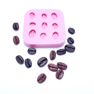 May include: Pink silicone mold with 12 cavities shaped like coffee beans. Scattered around the mold are 14 brown coffee beans.