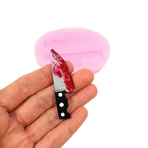 Peut inclure: Un petit couteau argenté avec un manche noir et des éclaboussures de peinture rouge sur la lame. Le couteau est tenu dans une main, et un moule en silicone rose est visible en arrière-plan.