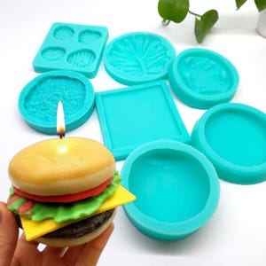 May include: A collection of turquoise silicone moulds in various shapes including squares, circles, and leaf patterns, alongside a lit hamburger-shaped candle with a tan bun, red tomato, green lettuce, yellow cheese, and a brown patty.