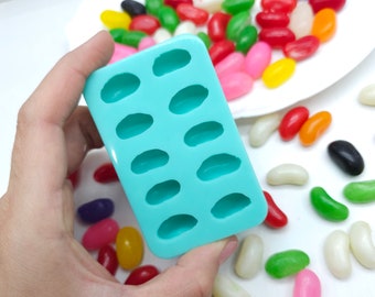 May include: A light blue silicone mold with 10 oval-shaped cavities. The mold is being held in a hand. There are colorful jelly beans scattered around the mold.