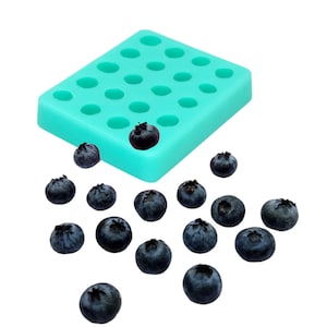 Peut inclure: Un moule rectangulaire en silicone bleu sarcelle clair doté de vingt cavités circulaires disposées en grille, accompagné d'un assortiment de myrtilles fraîches bleu foncé placées devant le moule sur un fond blanc uni.