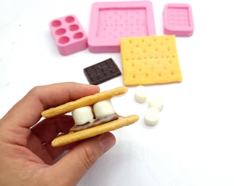 May include: A s'more made with two graham crackers, chocolate, and marshmallows. The s'more is being held in a hand. There are also pink silicone molds for making s'mores, a chocolate bar, and marshmallows in the background.