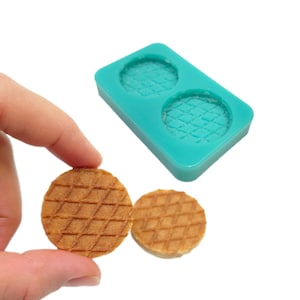 May include: A teal silicone mold with two circular waffle impressions, next to two small, round, golden-brown waffles. The waffles have a diamond pattern. The mold is rectangular and the waffles are held by a hand.