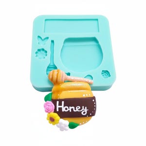 Puede incluir: Un molde de silicona azul claro con un diseño de tarro de miel. El tarro de miel es amarillo con una etiqueta marrón que dice "Honey". Un cucharón de miel de madera descansa encima. El molde también presenta una mariposa, una flor y una rosa.