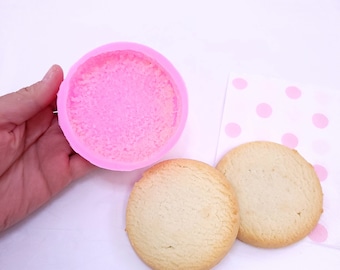 Puede incluir: Molde de silicona rosa para hacer galletas redondas, junto a dos galletas redondas sobre una superficie blanca con lunares rosas.