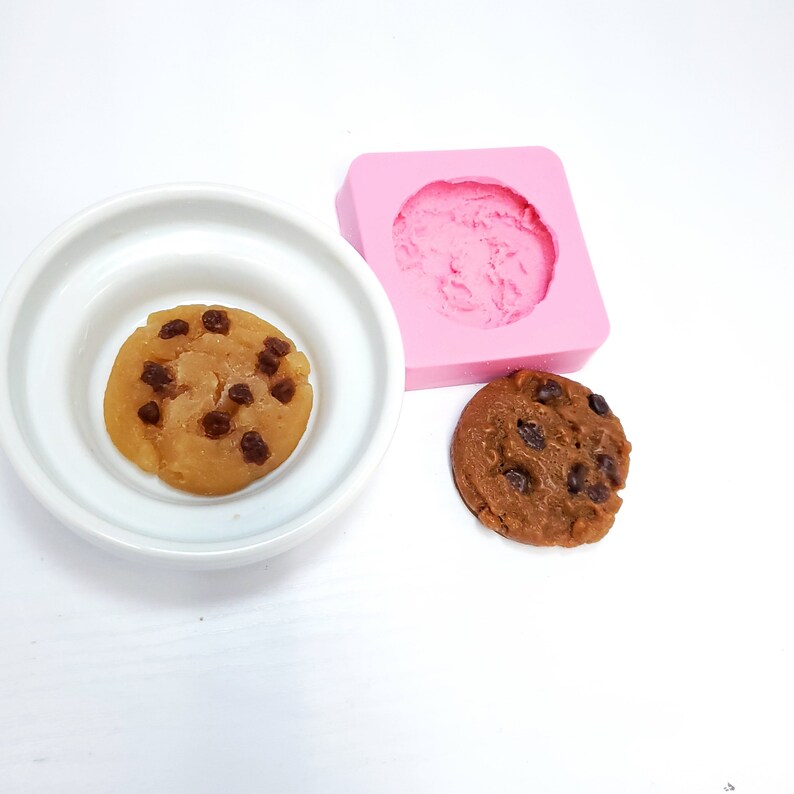 May include: A light brown cookie with dark chocolate chips sits in a white bowl next to a pink silicone mold and a finished cookie. The cookie is round and has a slightly textured surface.