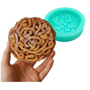 May include: A fried pastry shaped like a brain, held in a hand, next to a teal silicone mold in the shape of a brain. The pastry is golden brown with powdered sugar. The mold is round with a brain-like pattern.