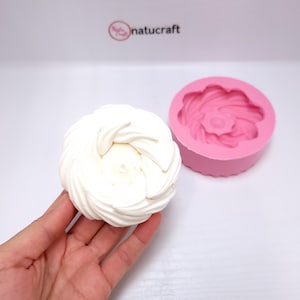 Op de afbeelding: Een wit, spiraalvormig dessert naast een roze siliconen mal. Het dessert wordt in een hand gehouden, wat het ingewikkelde ontwerp laat zien. De mal heeft een bloemachtig patroon. Het logo "natucraft" is zichtbaar op de achtergrond.