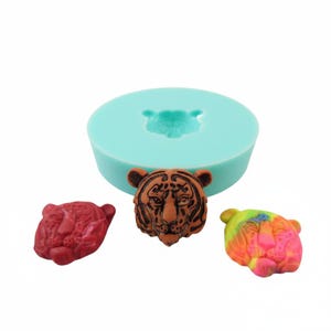 May include: A light blue silicone mould featuring a tiger head design, with three tiger head castings in red, brown, and multicoloured shades. The mould is round, and the castings are detailed.