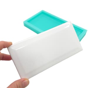 May include: A white rectangular tile held by a hand, with a glossy finish. A teal-colored silicone mold is in the background. The tile has beveled edges and a smooth surface, suggesting a craft or DIY project.