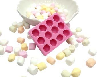 Può includere: Uno stampo in silicone rosa con 15 cavità circolari, circondato da marshmallow colorati. Sullo sfondo, una ciotola bianca con bordo smerlato piena di marshmallow. I marshmallow sono di vari colori pastello, tra cui rosa, giallo e bianco.