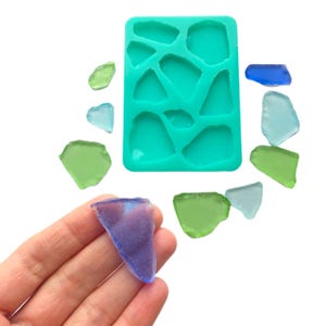 Puede incluir: Un molde de silicona turquesa con 10 cavidades de diferentes formas para hacer joyas de resina. El molde se muestra con varias piezas de vidrio transparente de diferentes colores y formas.