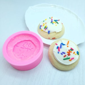Peut inclure: Moule en silicone rose pour faire des biscuits ronds avec un glaçage blanc et des pépites colorées. Deux biscuits sont présentés sur une assiette blanche.