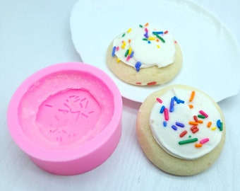 Puede incluir: Molde de silicona rosa para hacer galletas redondas con glaseado blanco y chispas de colores. Se muestran dos galletas en un plato blanco.