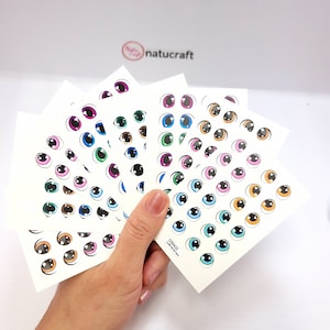 Puede incluir: Hojas de pegatinas de ojos en varios colores, incluyendo rosa, azul, verde y marrón. Cada hoja presenta múltiples pares de ojos de dibujos animados con contornos negros. El logotipo de Natucraft es visible en el fondo.