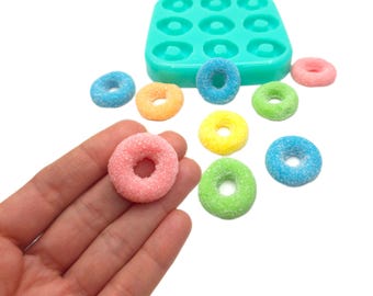 May include: A silicone mold with nine donut-shaped cavities, and a hand holding a pink donut with sprinkles. There are eight other donuts in various colors, including yellow, blue, green, and orange, scattered on a white surface.