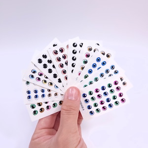 Puede incluir: Una mano sosteniendo varias hojas de pegatinas de ojos con varios colores y estilos. Las pegatinas presentan iris negros, marrones, azules, verdes y rosas. Las pegatinas están sobre un fondo blanco.