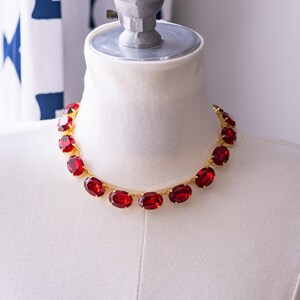 May include: A red rhinestone necklace with oval-shaped stones set in gold metal. The necklace is displayed on a white mannequin bust.