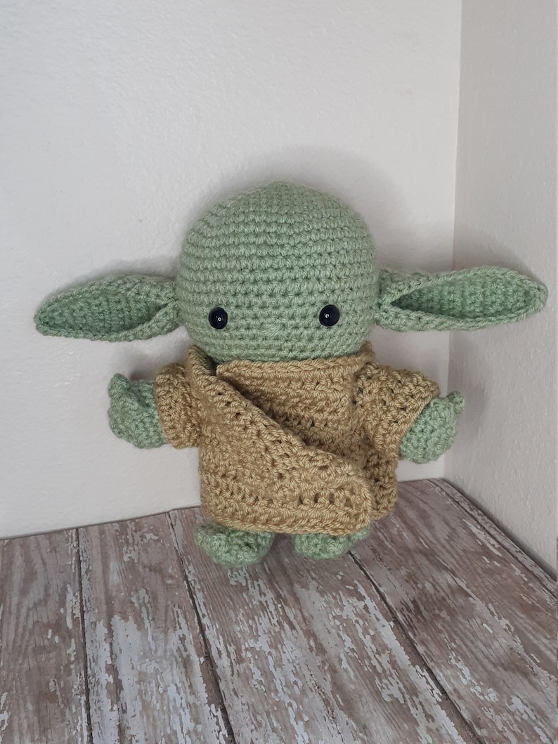 Puede incluir: Un juguete de peluche verde tejido a crochet con orejas grandes y un su&eacute;ter tejido a crochet marr&oacute;n. El juguete tiene ojos negros y una peque&ntilde;a boca.