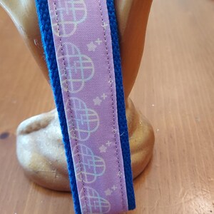 May include: A key fob with a silver-tone metal clasp and a blue and lavender strap. The lavender strap features a repeating pattern of a globe-like design. The key fob is a practical accessory for organizing keys.