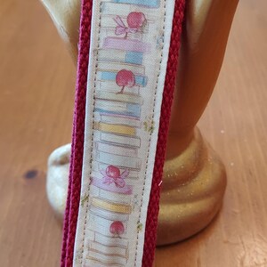 May include: A burgundy key fob with a silver-colored metal clasp. The fabric features a repeating pattern of stacked books, cherries, and bows in pastel colors. The key fob is held by a gold-colored hand-shaped object.