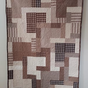 May include: A brown and white patchwork quilt with a variety of patterns, including plaid, stripes, and houndstooth. The quilt is hanging on a wall with a wooden rod.