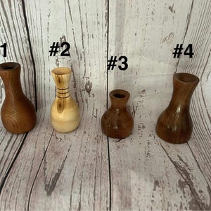 May include: Four small, handcrafted wooden vases of varying shapes and colors are displayed. The vases are numbered 1 through 4. The wood tones range from light tan to dark brown. The background is a light wood grain.