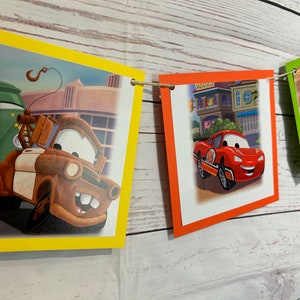 Puede incluir: Una pancarta colorida con im&aacute;genes de coches de dibujos animados. La pancarta est&aacute; hecha de papel y tiene una cuerda que la atraviesa. Los coches son rojos, verdes, marrones y azules. La pancarta es perfecta para una fiesta de cumplea&ntilde;os u otra celebraci&oacute;n.