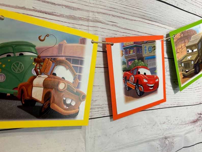 Puede incluir: Una pancarta colorida con im&aacute;genes de coches de dibujos animados. La pancarta est&aacute; hecha de papel y tiene una cuerda que la atraviesa. Los coches son rojos, verdes, marrones y azules. La pancarta es perfecta para una fiesta de cumplea&ntilde;os u otra celebraci&oacute;n.