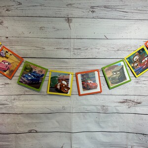 Puede incluir: Un bander&iacute;n colorido con tem&aacute;tica de Cars con im&aacute;genes de diferentes personajes de la pel&iacute;cula. El bander&iacute;n est&aacute; hecho de cart&oacute;n y tiene una cuerda que lo atraviesa. El bander&iacute;n est&aacute; colgado en una pared de madera blanca.