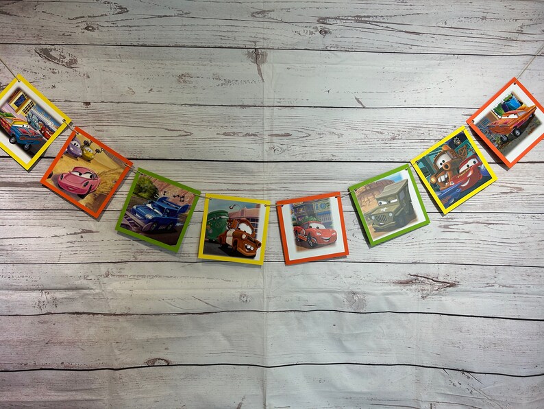 Puede incluir: Un bander&iacute;n colorido con tem&aacute;tica de Cars con im&aacute;genes de diferentes personajes de la pel&iacute;cula. El bander&iacute;n est&aacute; hecho de cart&oacute;n y tiene una cuerda que lo atraviesa. El bander&iacute;n est&aacute; colgado en una pared de madera blanca.