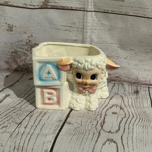 May include: A cream-colored ceramic planter shaped like a lamb sitting next to two stacked alphabet blocks. The top block features a blue letter A, and the bottom block features a pink letter B. The lamb has large black eyes and a pink bow.