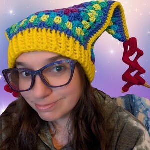 May include: A colorful, handmade crochet hat with a long, pointed end. The hat features a yellow brim, a top section with blue, green, and purple squares, and a red, zig-zag shaped embellishment. The hat is worn by a person wearing glasses.